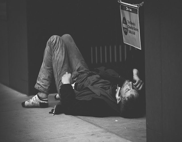 Disheveled person lying on their back on a public place with their hand raised to their face