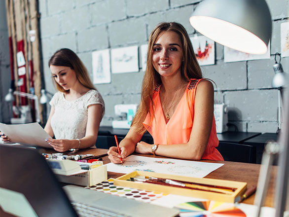 Two women sit at a shared art studio desk, working on watercolor and pencil drawings. The woman in the foreground, wearing a bright orange top, smiles at the camera while holding a red pencil. Art supplies, a color wheel, and a laptop are spread out on the desk under warm studio lighting.