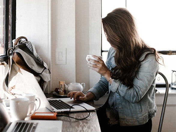 A woman with long hair sits at a window-side counter in a cozy café, holding a cup and working on a laptop. She wears a light denim jacket and has a backpack nearby. The table is scattered with another laptop, mugs, a teapot, and a notebook.