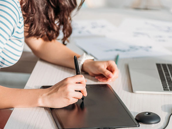 A person wearing a striped shirt uses a digital drawing tablet at a desk, surrounded by sketch papers and a laptop. They are working on illustrations, with one hand holding a stylus and the other resting on the tablet. The workspace is bright and creative.