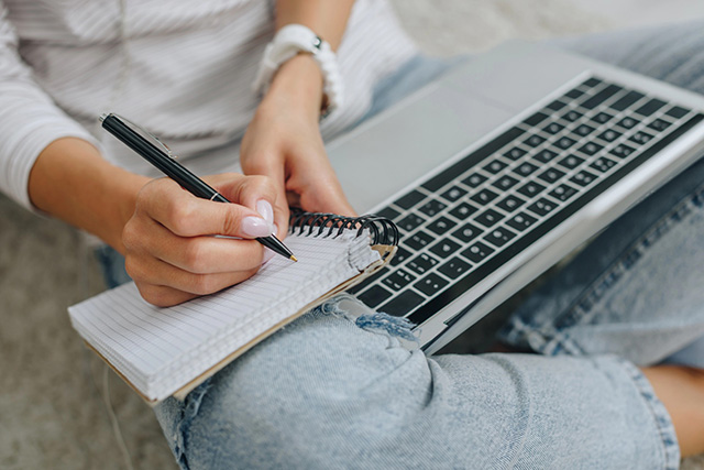 Close-up of hand writing in a notebook resting on a laptop