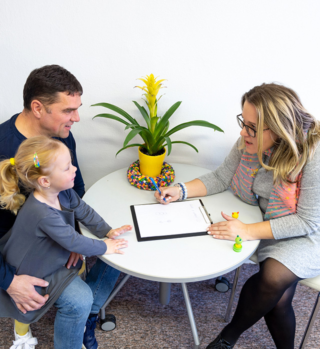 Little girl seated a table with two adults, one of whom is writing on clipboard