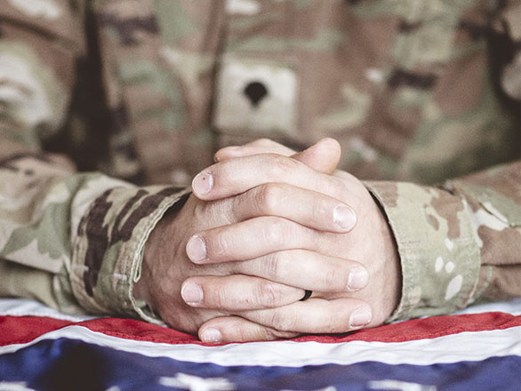 A person wearing a military uniform with camouflage rests their hands on an American flag.
