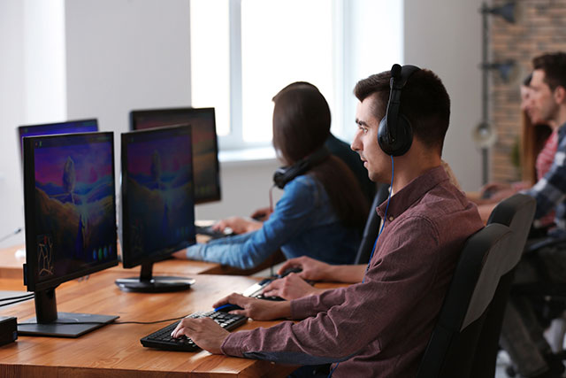 Several people wearing headsets playing video games at desktop computers in a bright room.