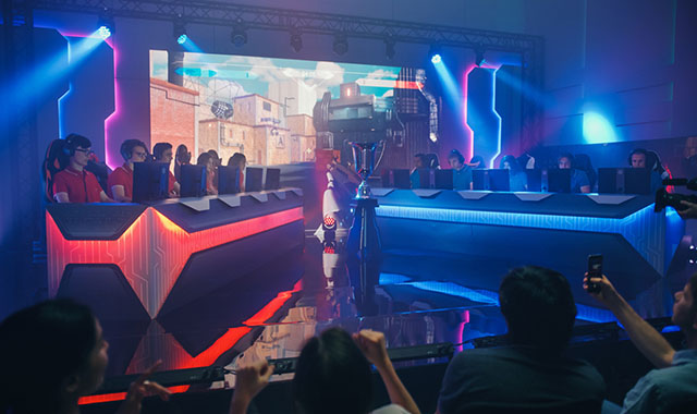 Two esports teams compete on a lit stage with a large screen displaying gameplay, as audience members watch and record.