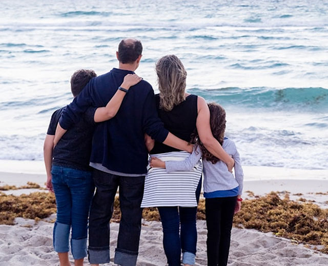 Family of four seen from behind, embracing while standing on a sandy beach, looking out on the water