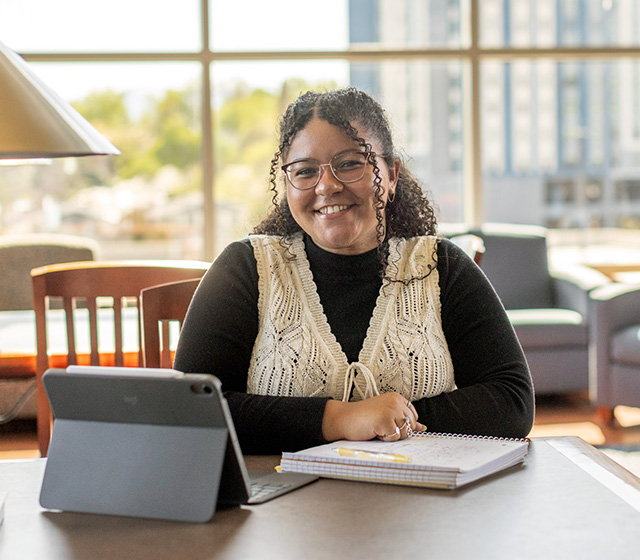 Woman seated a table, writing in a notebook while smiling at the camera