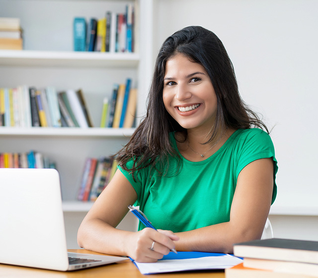 Seated young woman writing in a notebook while smiling at the camera