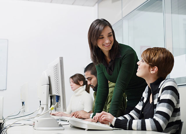 Woman leaning on a table, smiling at another woman working on a computer