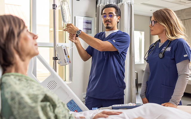 Two nursing students in navy scrubs attend to a patient at her bedside, with one adjusting an IV pump while the other observes.