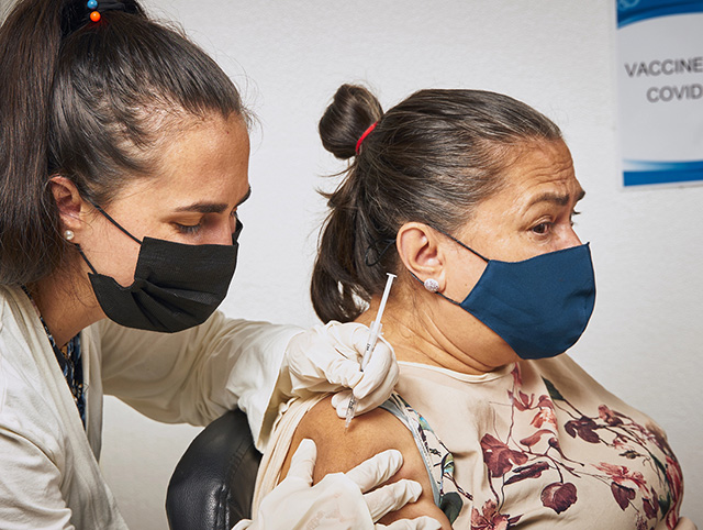 Masked woman giving another masked woman an injection in the shoulder