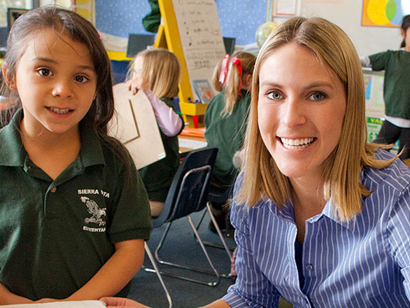 A young girl in a green “Sierra Vista Elementary” polo shirt smiles next to a teacher in a blue striped shirt, seated in a colorful classroom.