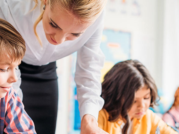 A teacher in a white blouse leans over to assist a young boy, while another student focuses on her work in the background.