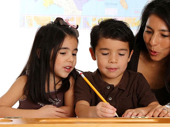 A young boy and girl sit at a desk working together on a writing activity, with the boy holding a pencil. An adult woman, possibly their teacher, leans in to guide and encourage them. A world map is visible on the wall in the background.