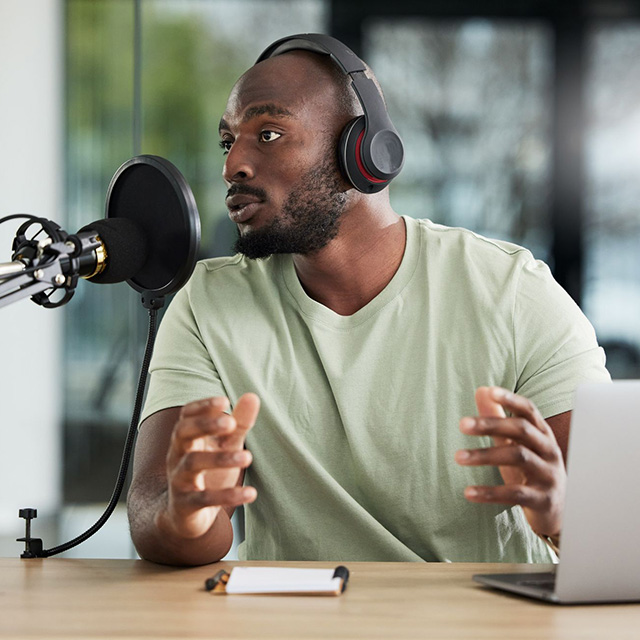 Man wearing headphones, seated at a table in front of a laptop, speaking into a microphone