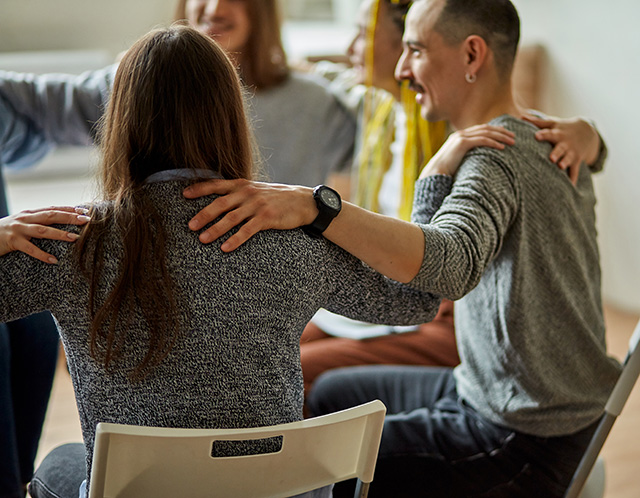 Members of a support group sit in a circle, placing their arms around one another's shoulders.