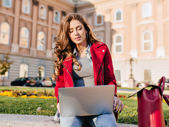A young woman sits on a stone bench outside a historic university building, working on a laptop. She wears a red jacket and jeans, with a red bag beside her. Flowers bloom in the grass behind her on a sunny day.