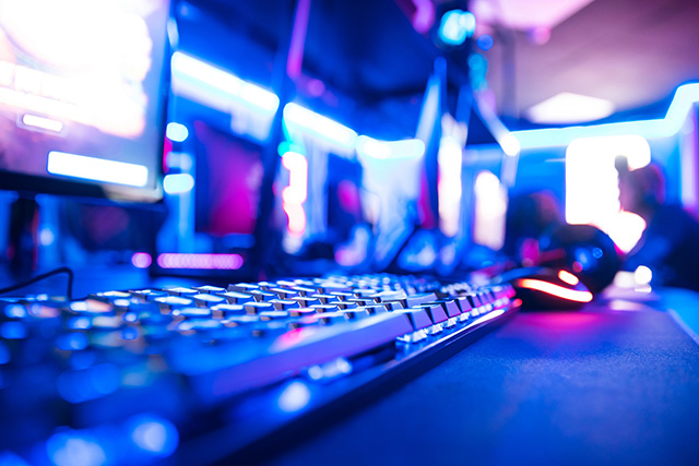 Two esports teams compete on a lit stage with a large screen displaying gameplay, as audience members watch and record.