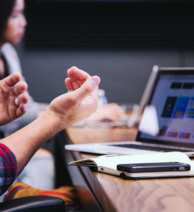 Close-up of hands gesturing at a table with open laptops, a notebook and a smartphone.