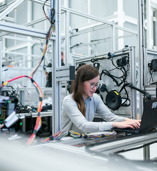 A woman wearing glasses works at a laptop in a technical lab environment surrounded by electronic equipment, wiring, and metal framework.