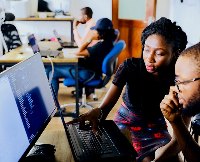 Two students collaborate at a laptop computer displaying code, in an open workspace.