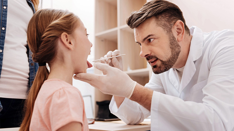 Doctor in white scrubs using a tongue depressor to examine a young girl