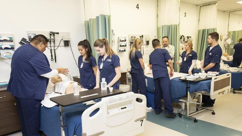 Two groups of nursing students practice on dummy patients in two separate beds in a hospital setting.