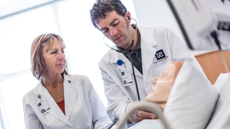 A nursing student wears a stethoscope and practices on a patient dummy while an instructor looks on.