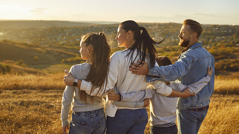 Family of four seen from behind, embracing while looking into a semi-arid landscape illuminated in late-afternoon light