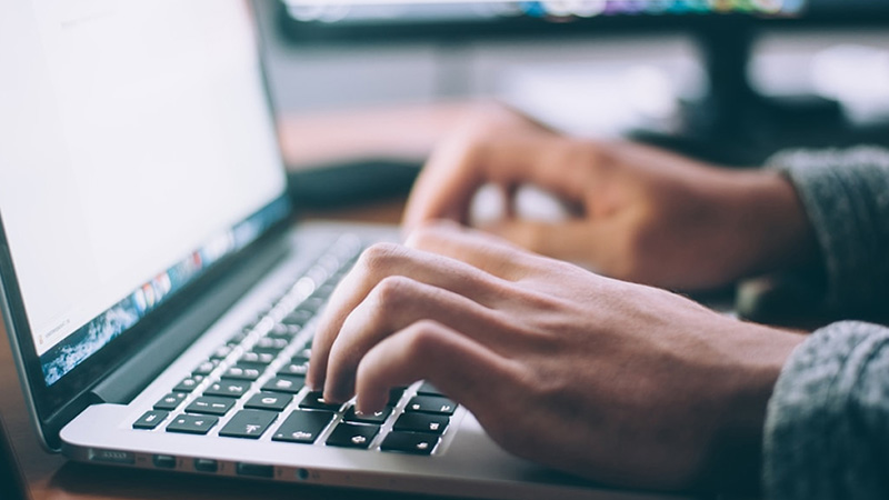 Close-up of a person's hands typing on a laptop keyboard, with a second monitor visible in the blurred background.