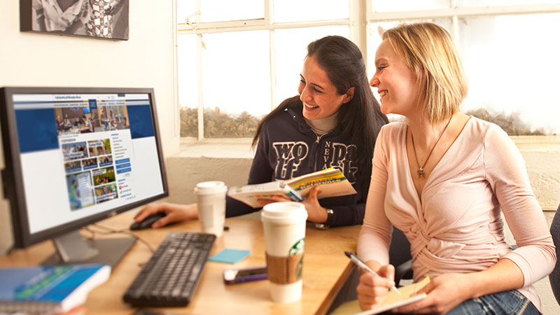 Two students look at a computer. Two cups of coffee and a notebook are on the table in front of the computer.